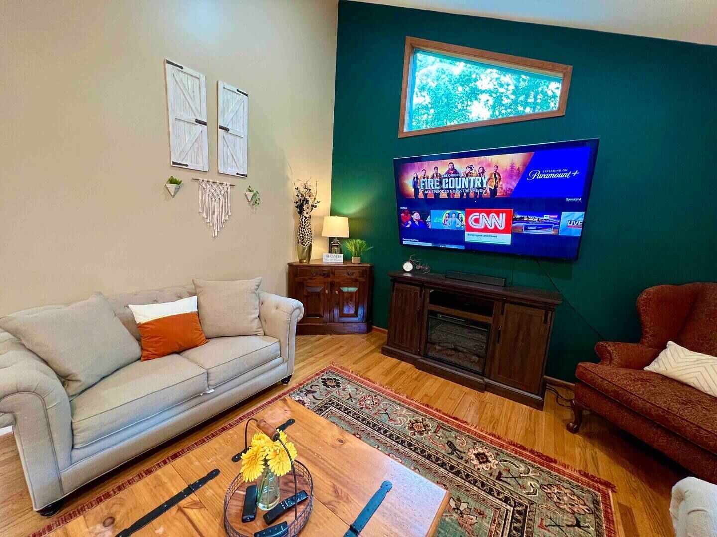 A living room with a grey sofa, a red armchair, a coffee table with a vase of flowers, a television mounted on the wall, and a framed picture above the TV.