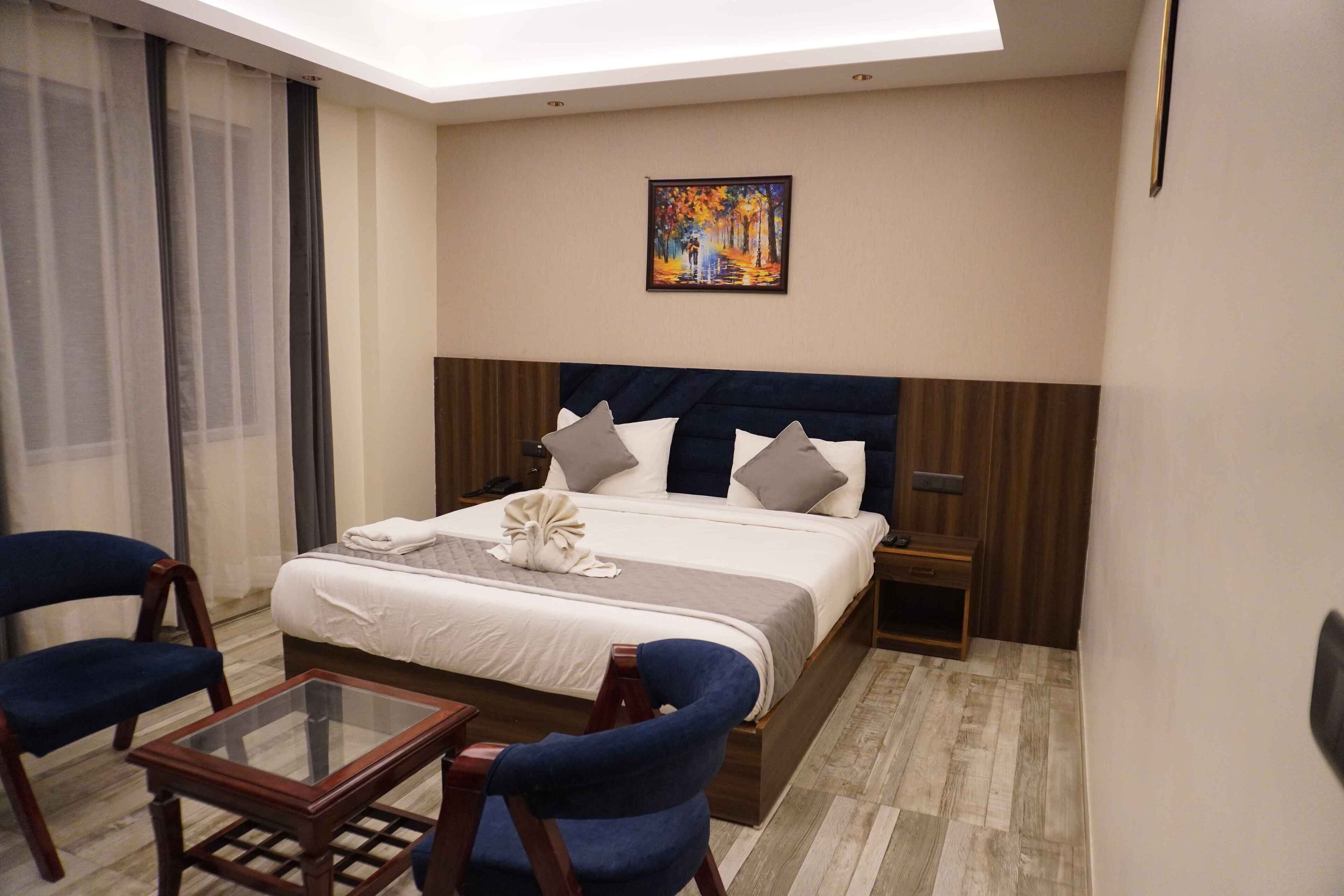 A hotel room with a bed, two chairs, a glass table, a painting on the wall, and a nightstand.