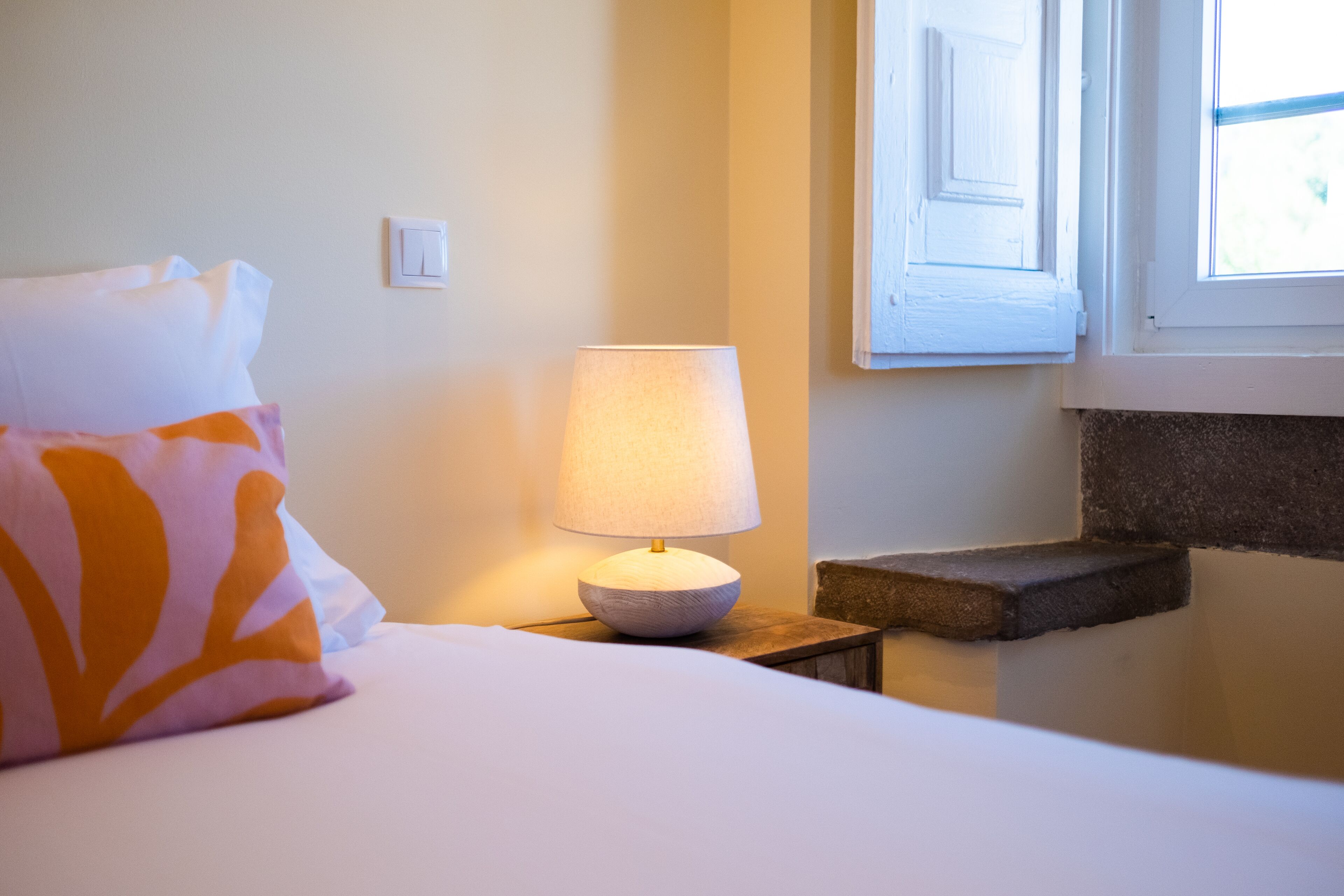 Una cama bien hecha con un almohadón blanco y naranja, una lámpara de noche y una ventana con contraventanas blancas.