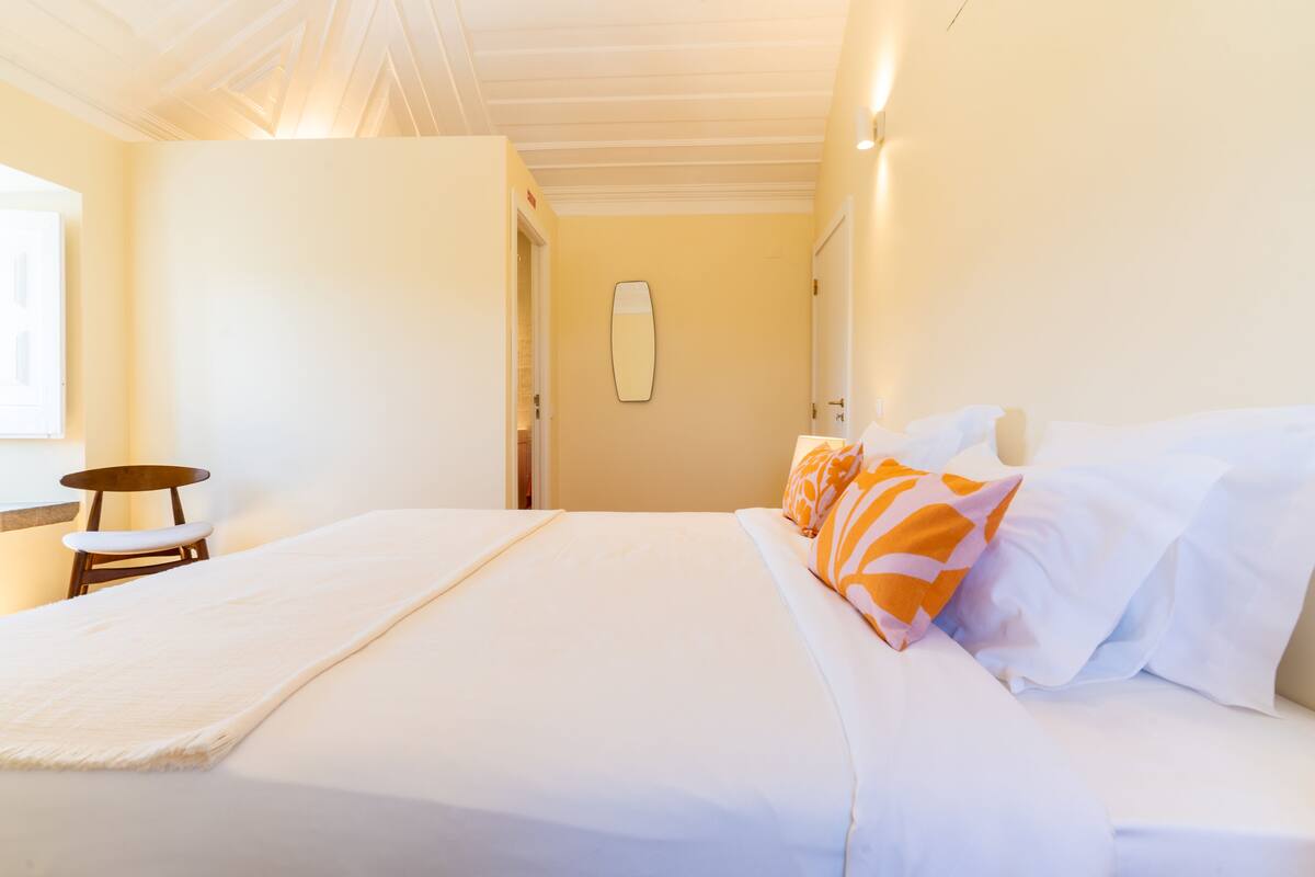 Una cama bien tendida con almohadas blancas y naranjas, una silla de madera y una mesita.