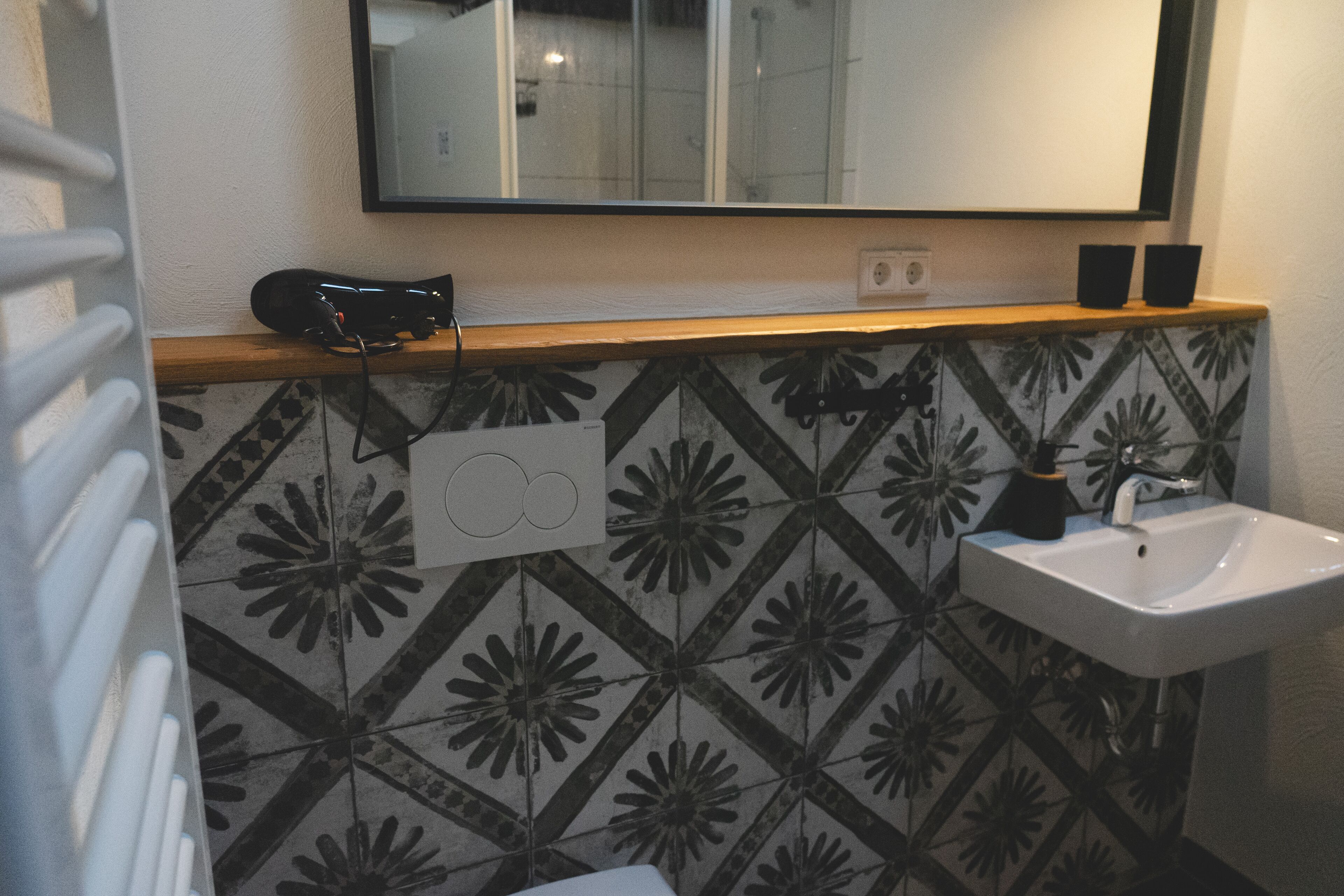 Une salle de bain avec un mur recouvert de carreaux décoratifs formant un motif géométrique, un lavabo blanc et un miroir au-dessus.