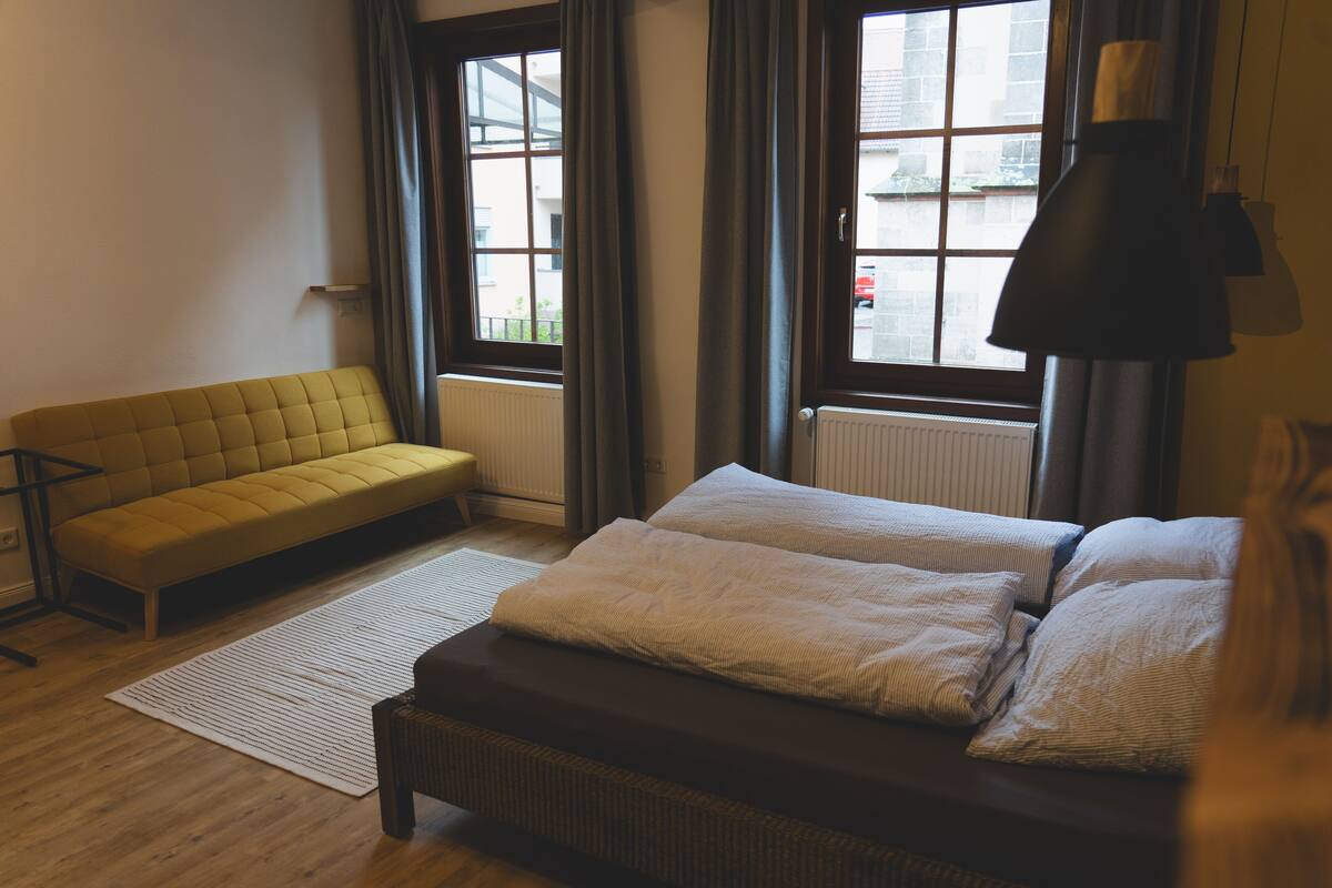 A room with a sofa, a bed with two pillows, a floor lamp, and a view through a window.