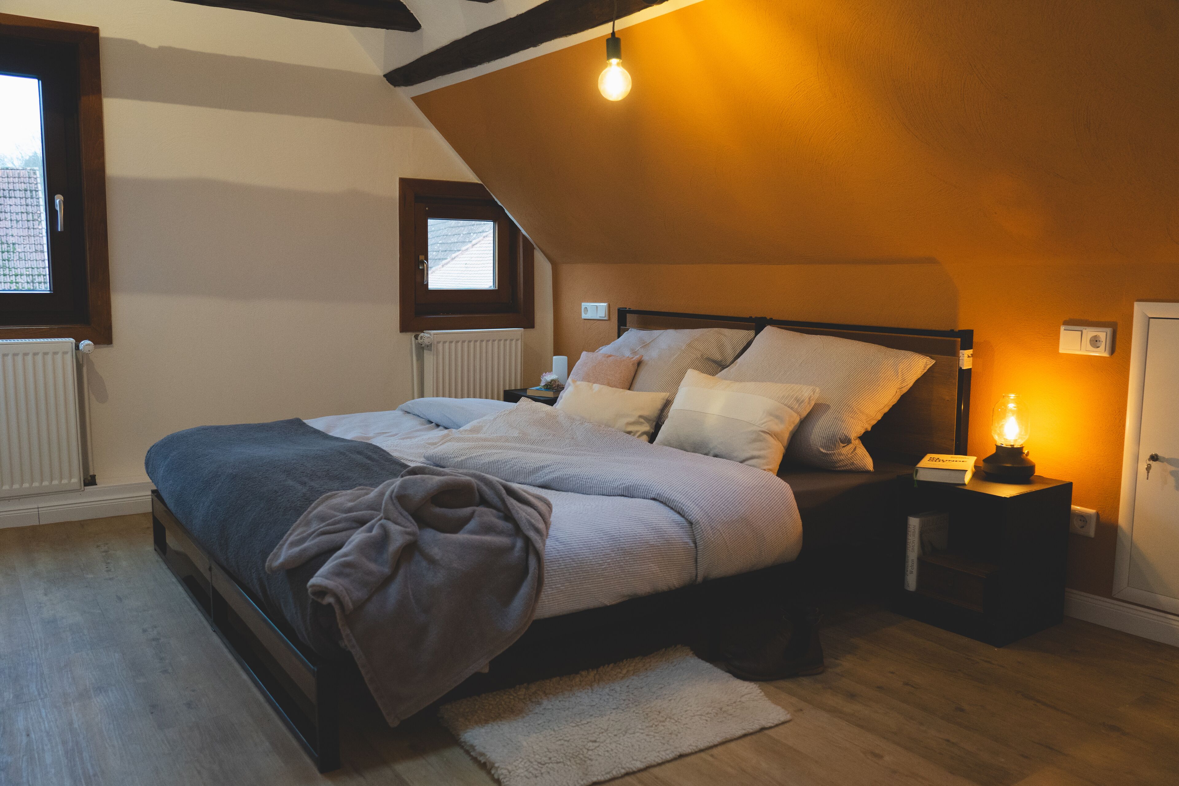 A bedroom with a sloped ceiling, a bed with a dark headboard, a bedside table with a lamp, and a window with a view.