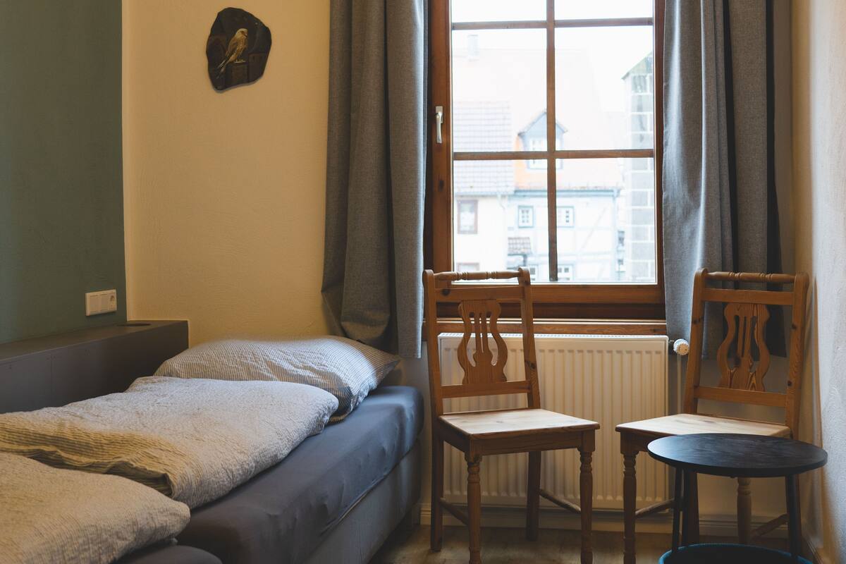 A small, cozy room with a bed, two wooden chairs, a small table, and a window with curtains.