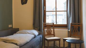 A small, cozy room with a bed, two wooden chairs, a small table, and a window with curtains.