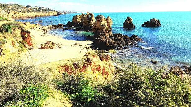 A coastal landscape with rocky formations, clear blue water, and a sandy beach.