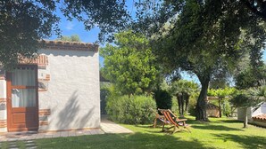 Uma casa com fachada branca, porta de madeira e entrada com azulejos. Há um jardim com gramado, cadeira de balanço de madeira e várias árvores.