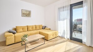 A modern living room with a mustard-colored sofa, a small wooden coffee table, and a cityscape view through a large window with sheer curtains. - Ferienwohnung Tamar - 6 Personen, 2 Schlafzimmer, Balkon (Umag)
