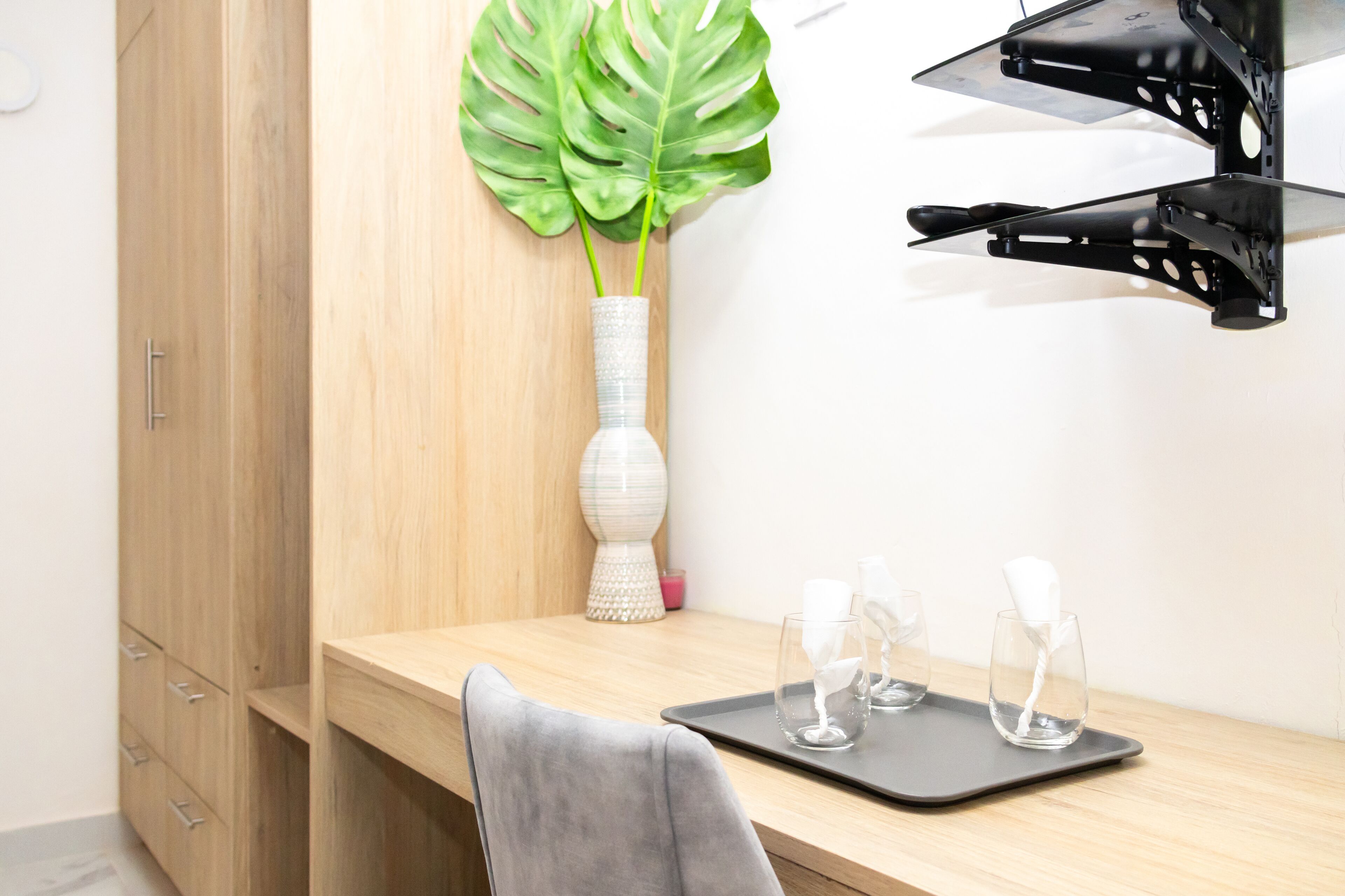 Quarto moderno com escrivaninha de madeira, um vaso branco com planta verde e bandeja preta com copos e guardanapo.