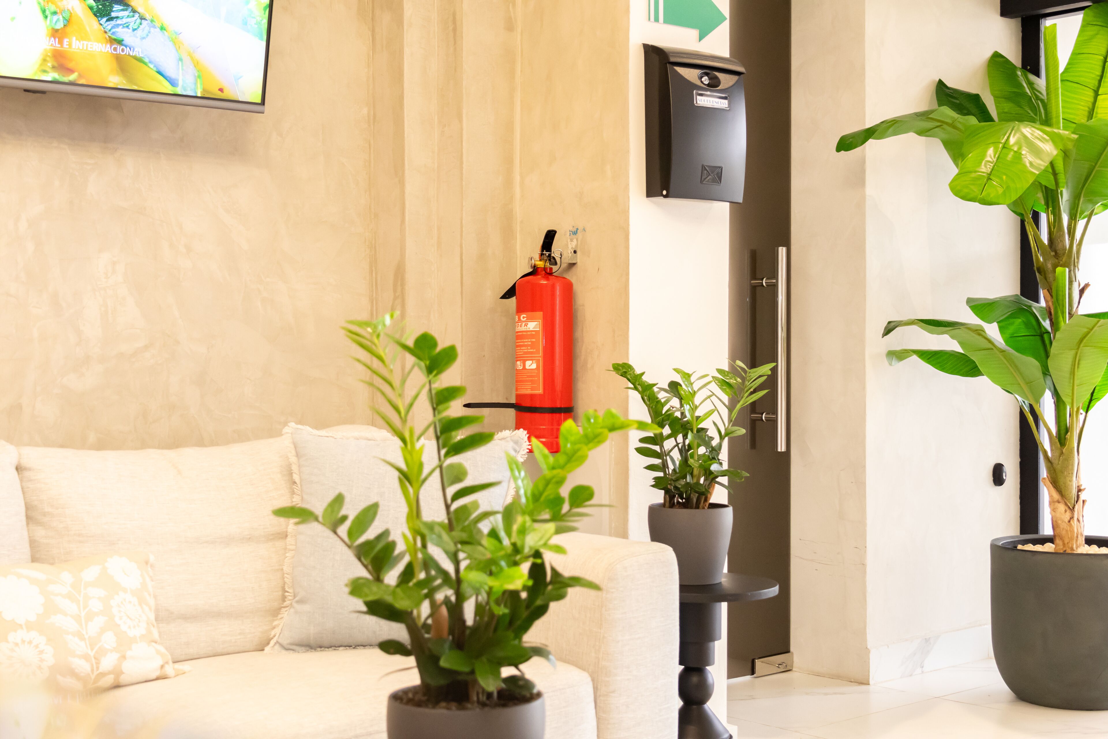 A modern hotel lobby with a beige sofa, potted plants, a fire extinguisher, and a digital screen displaying a colorful image.