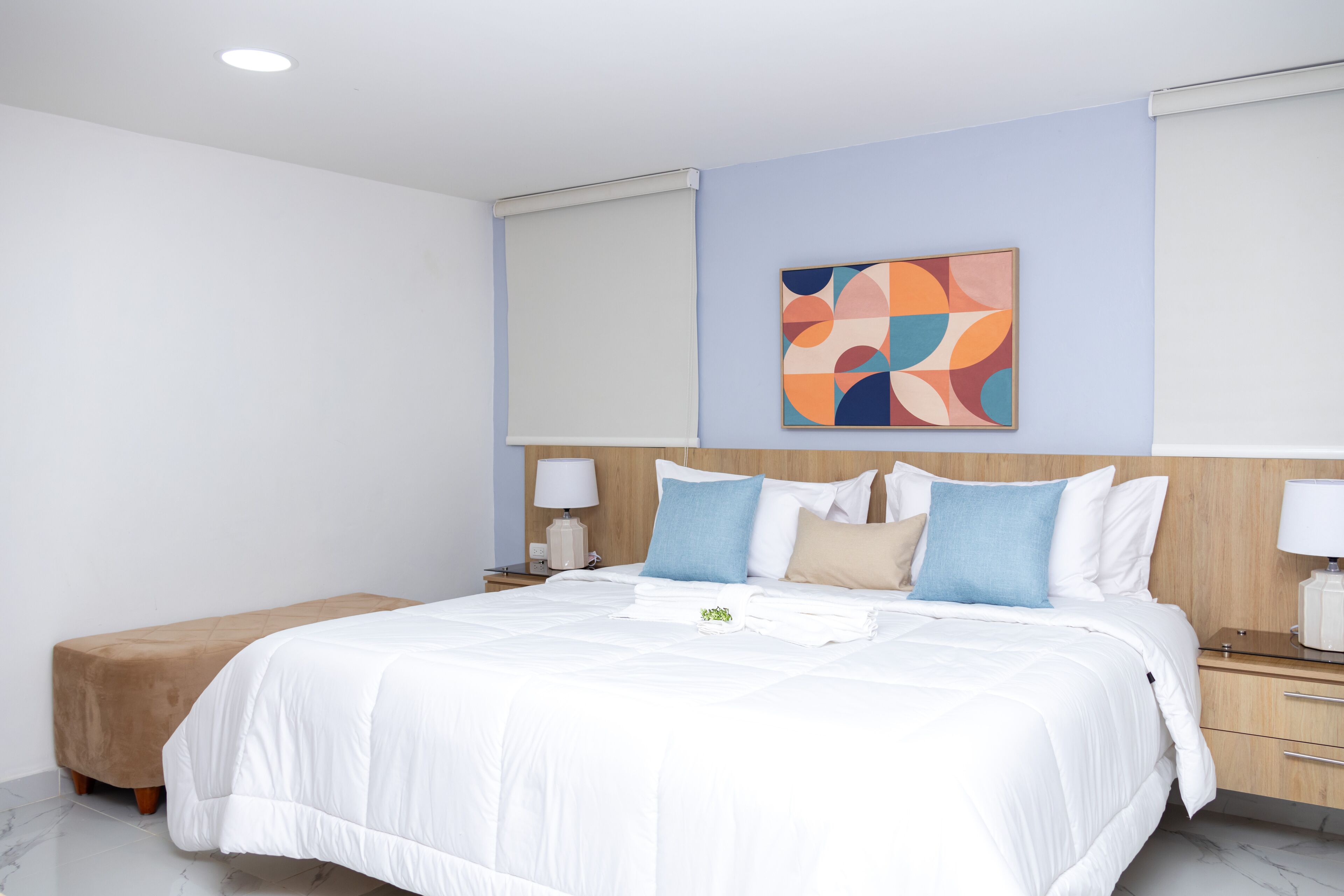 Een moderne hotelkamer met een groot bed, een houten hoofdeinde, een klein nachtkastje met een lamp en een kleurrijk abstract schilderij aan de muur.