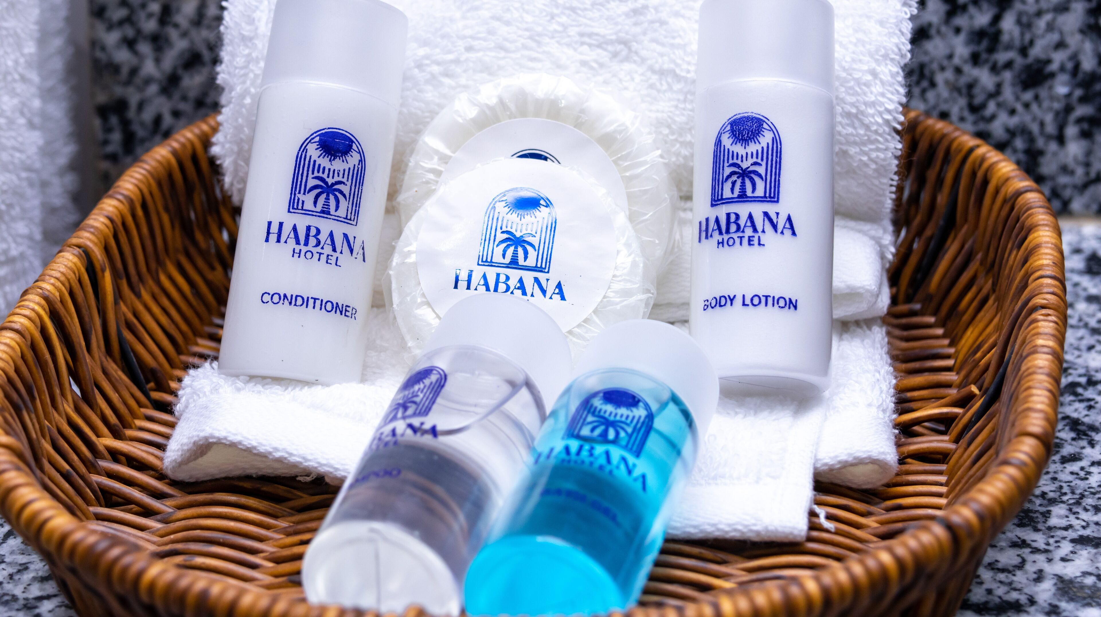 Un panier en osier contenant trois flacons de lotion pour le corps de la marque Habana Hotel, un flacon d’après-shampoing et un flacon bleu, le tout posé sur une serviette blanche.