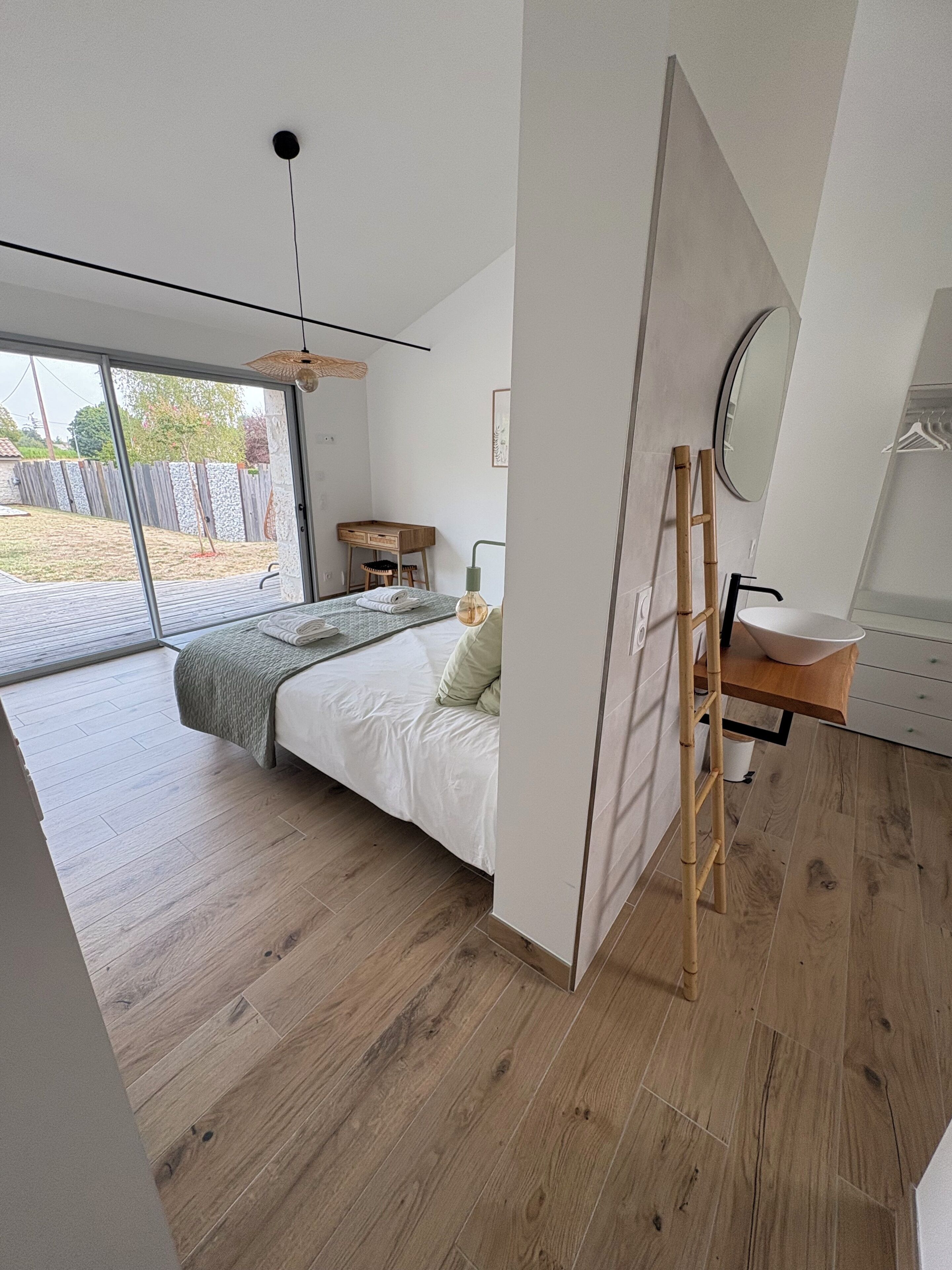 A modern bedroom with a bed, a wooden desk, a mirror, and a bathroom with a sink.