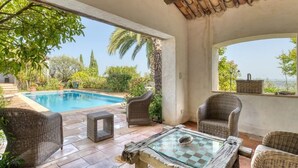 An outdoor patio with wicker furniture, a glass table, and a view of a swimming pool and greenery. - Peaceful villa (Tourrettes-sur-Loup)