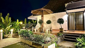 Patio esterno ben illuminato con zona salotto coperta, piante in vaso e una scalinata che conduce a un edificio.