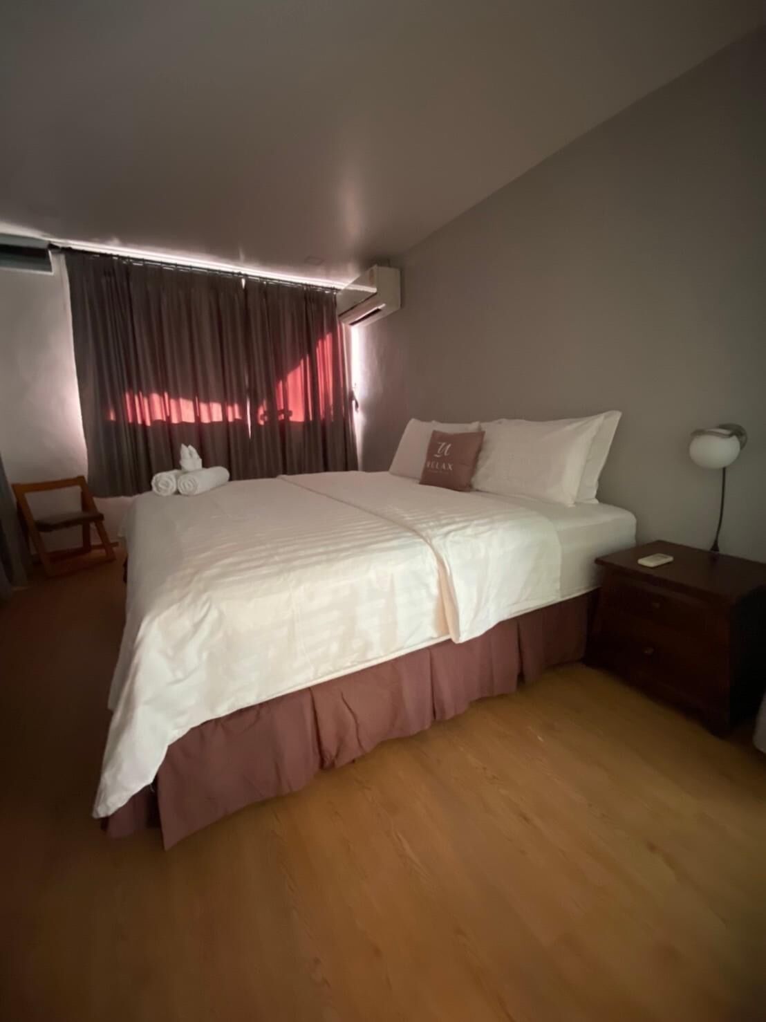 A neatly made bed with a white and pink bedspread, a wooden nightstand with a lamp, and a chair with a towel.
