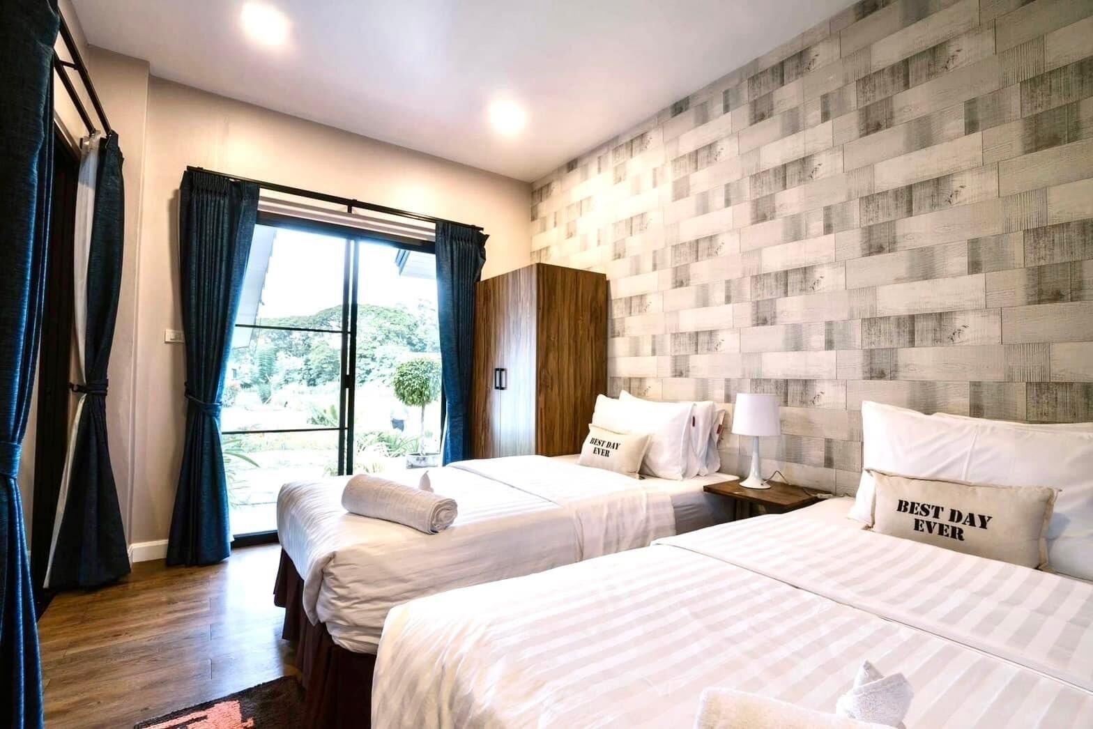 Quarto de hotel com duas camas, uma janela grande com cortinas, uma cabeceira de madeira e vista para o verde lá fora.