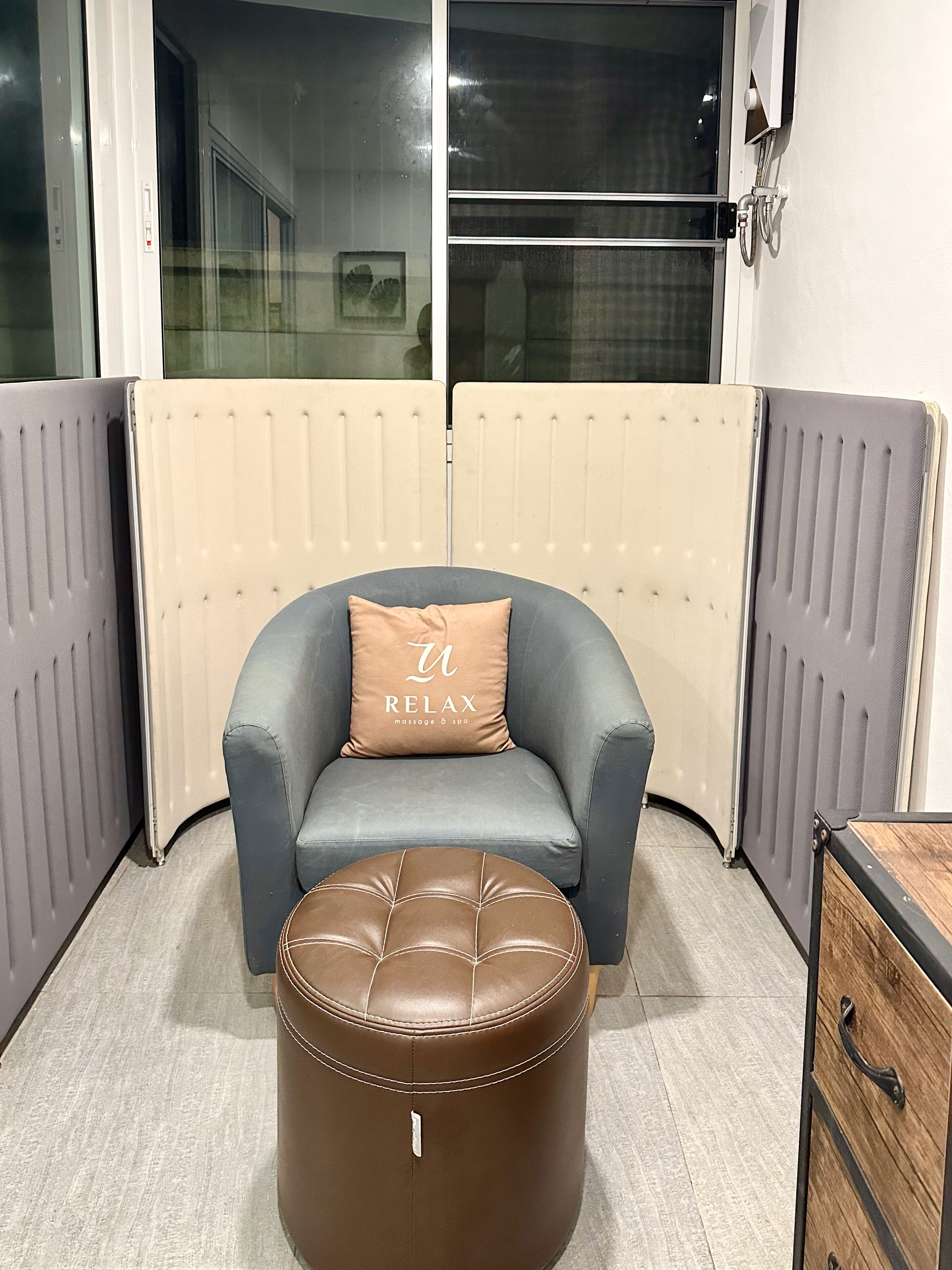 Ein Eckzimmer mit grauem Sessel, braunem Hocker und einem Holzschrank.