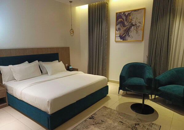 Een hotelkamer met een groot bed, twee blauwgroene fauteuils, een klein rond tafeltje, een schilderij aan de muur en een telefoon op het nachtkastje.