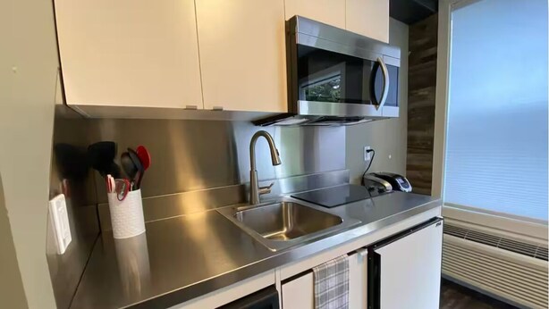 A modern kitchen with stainless steel appliances, a dishwasher, and a window with blinds. - Central Farmhouse-Style Micro Apartment (Lethbridge)