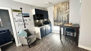Una cocina moderna con microondas, lavamanos y armarios oscuros. Hay un cartel de bienvenida en la pared y un comedor con mesa y sillas.