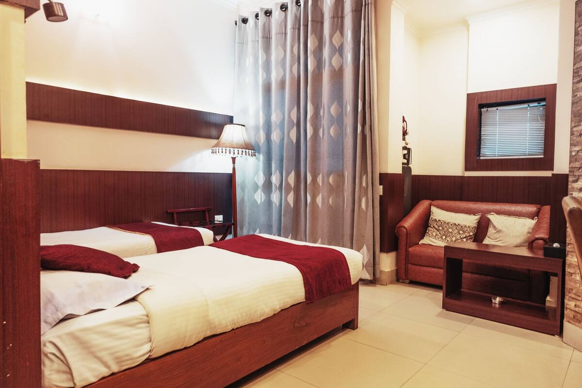 Une chambre d’hôtel avec deux lits en bois, un canapé rouge, une petite table et une fenêtre avec des stores.