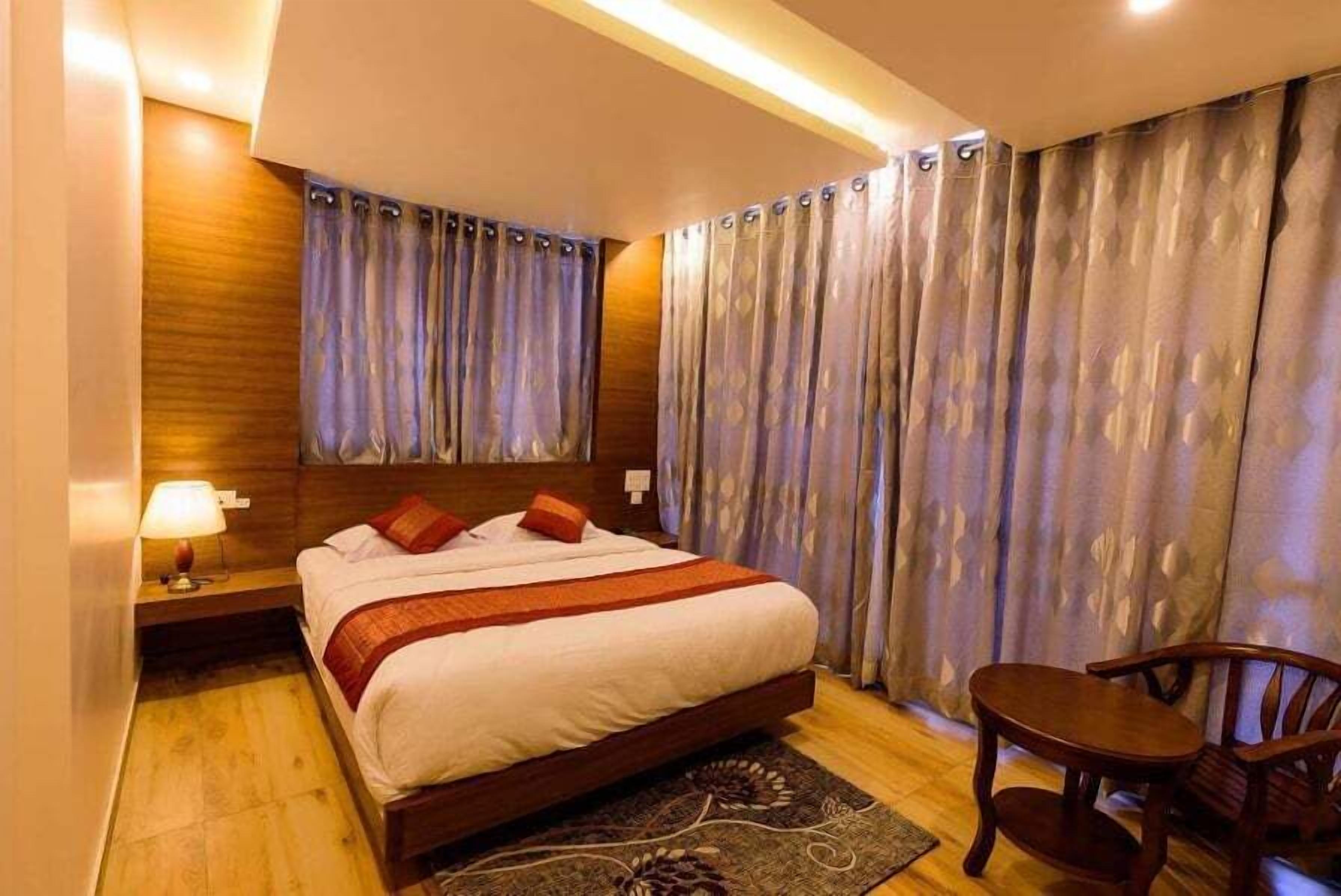 A hotel room with a large bed, wooden headboard, a nightstand with a lamp, a wooden chair, and a window with patterned curtains.