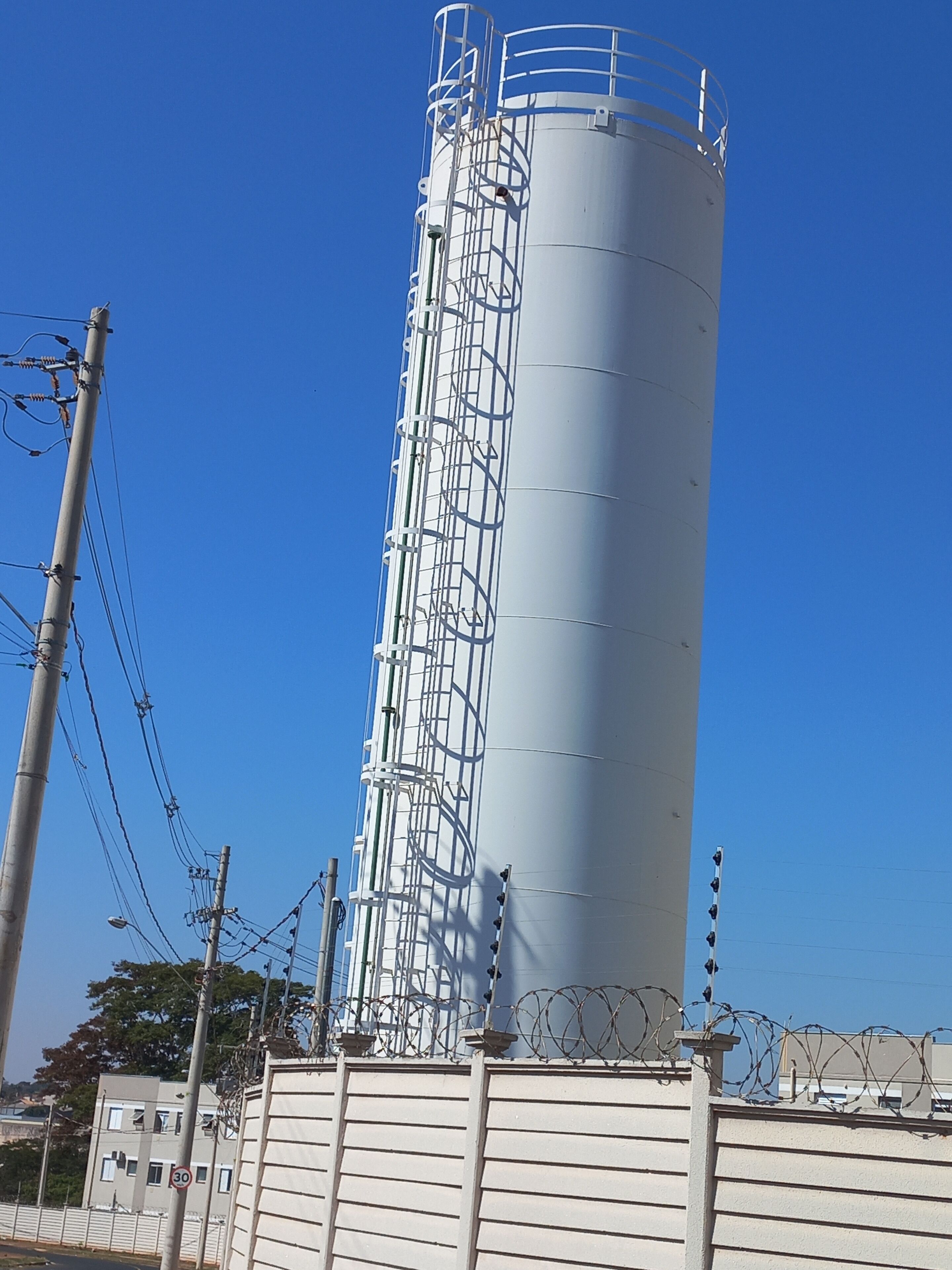 Un grand réservoir cylindrique servant de silo, doté d’une structure métallique et entouré d’une clôture surmontée de fil barbelé.
