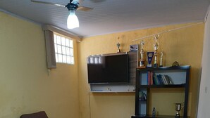A living room with a sectional sofa, a flat-screen TV mounted on the wall, a bookshelf with trophies, and a ceiling fan.