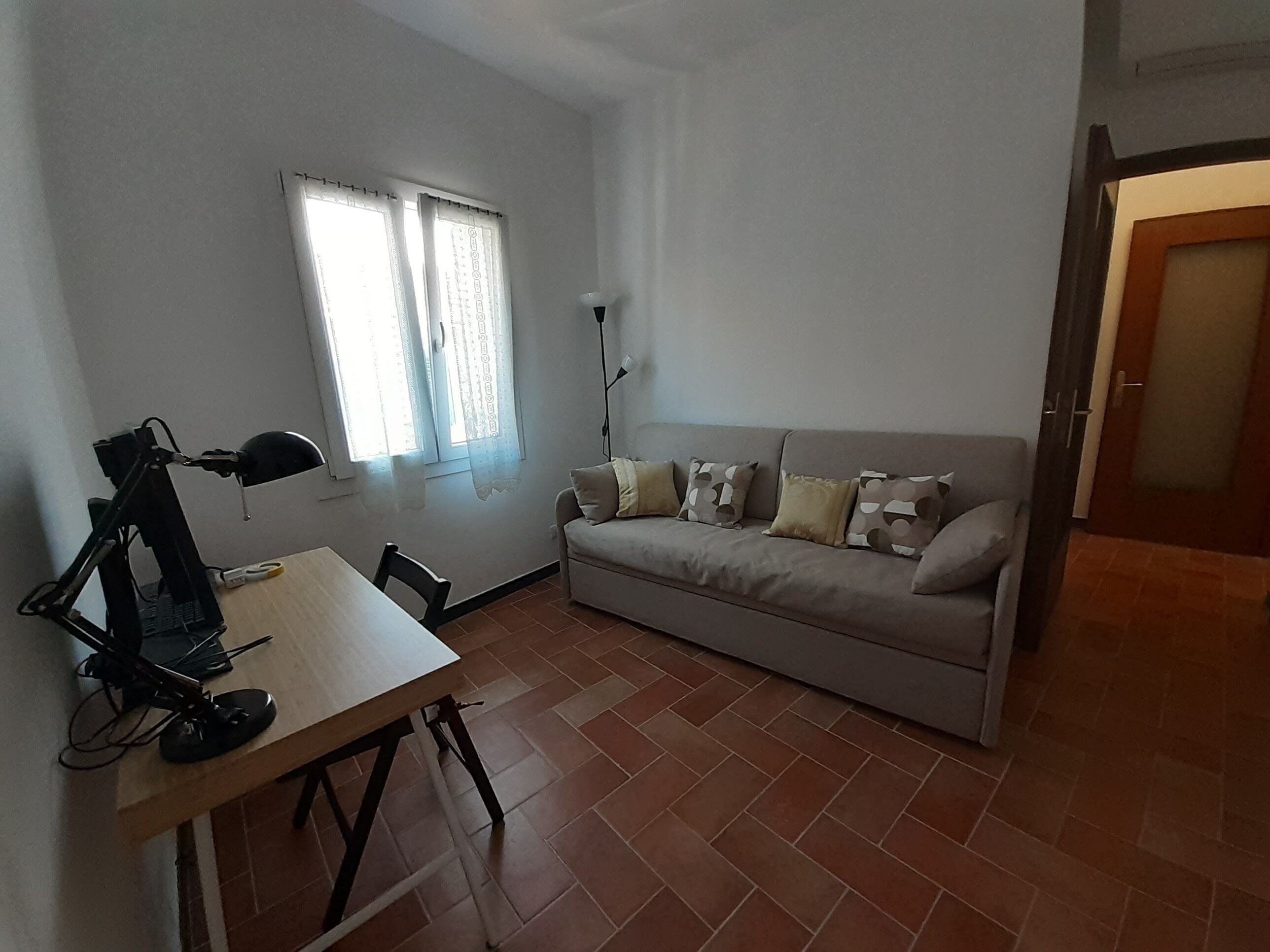 Une petite pièce bien éclairée, avec un canapé, une table équipée d’un ordinateur et une fenêtre avec des rideaux.