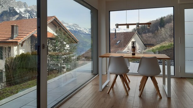 A modern room with large glass doors leading to a balcony, wooden flooring, and a dining table with chairs. - Apartment Panorama Walensee (Glarus Nord)