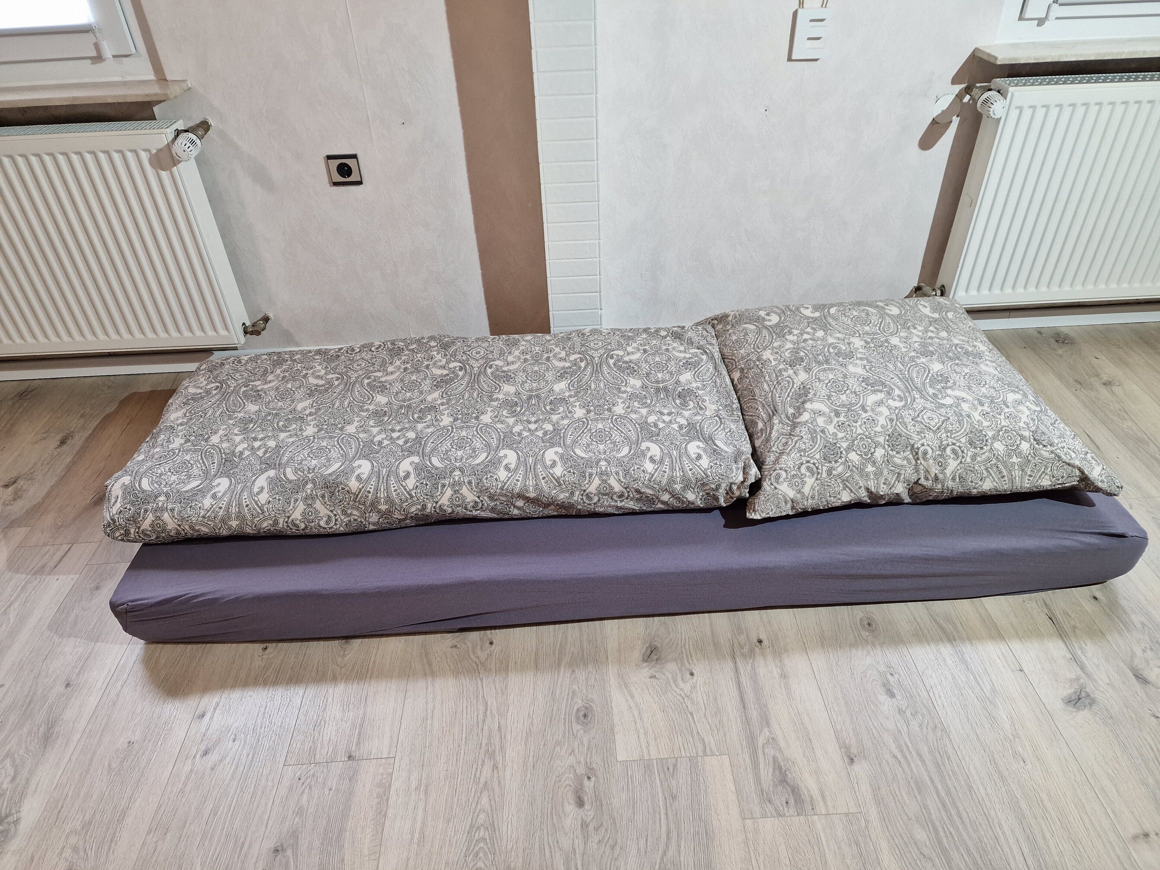 A bed with patterned pillows and a dark mattress in a room with wooden flooring and white walls.