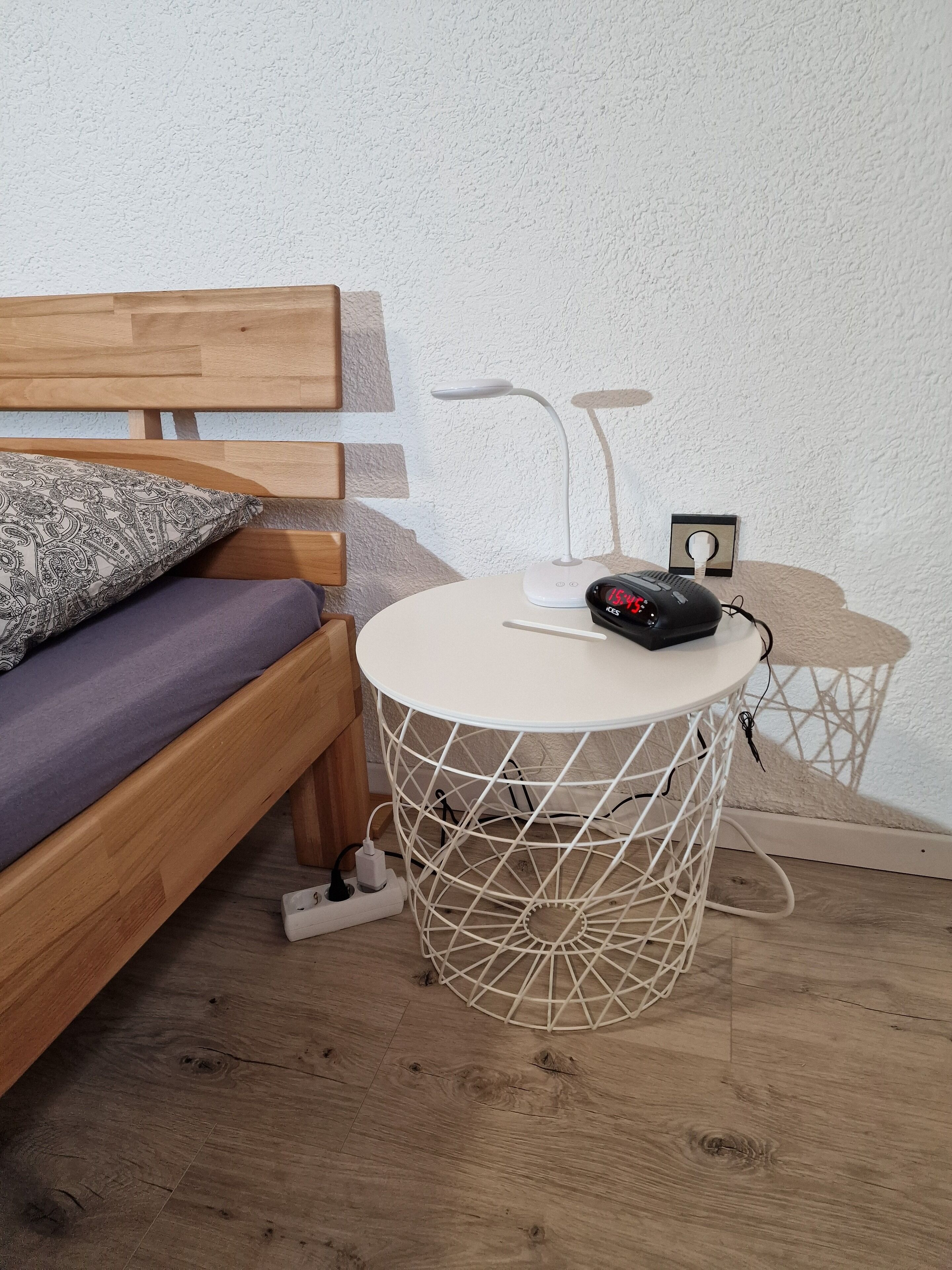 Un lit en bois avec un oreiller à motifs, une petite table ronde blanche avec une multiprise et une lampe.
