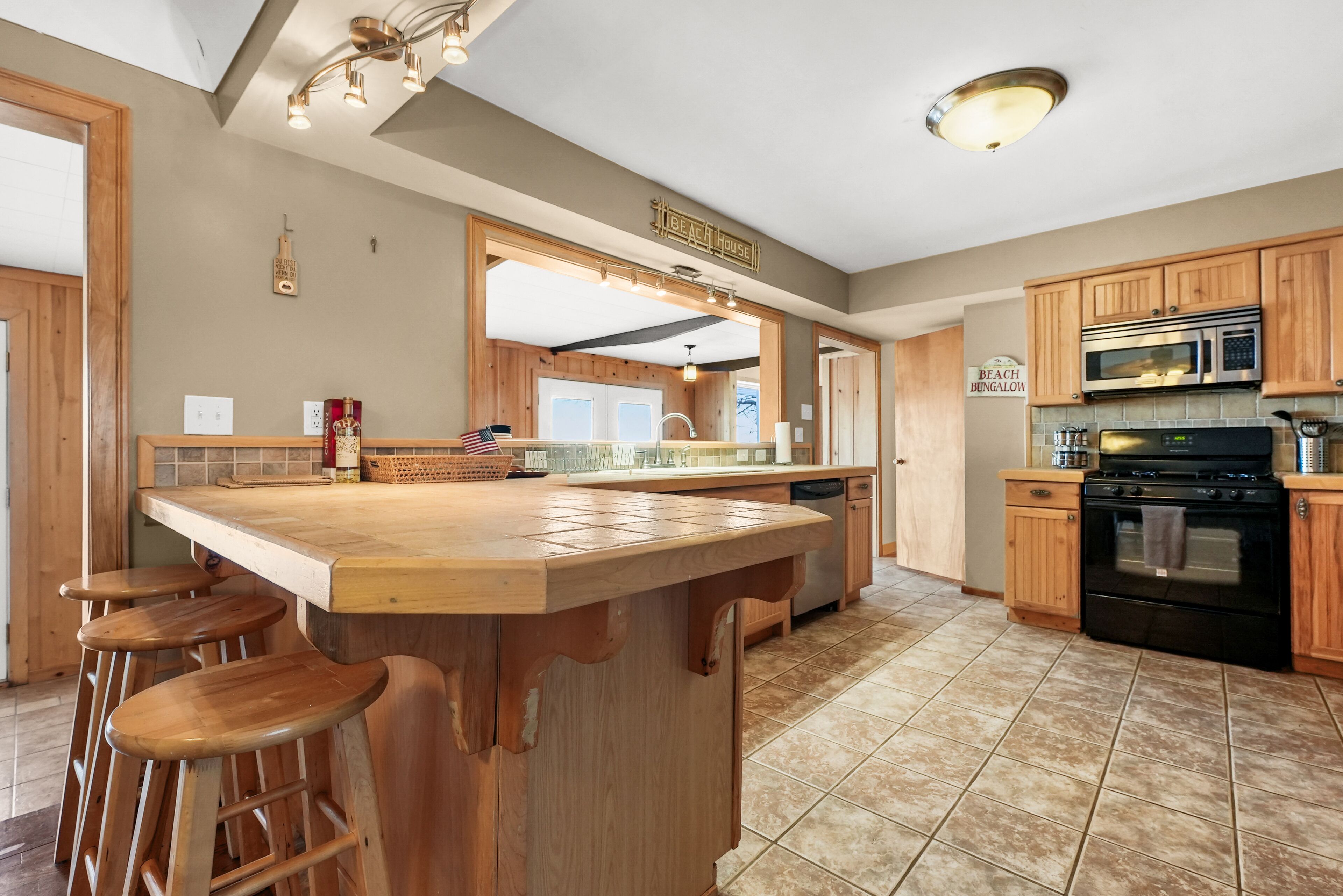 A kitchen with a large island, wooden bar stools, a microwave, oven, and a sign that reads "Beach House Bungalow".