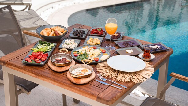 Mesa de jantar com diversos pratos e bebidas, montada junto a uma piscina.