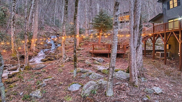 Una cabaña con terraza de madera y cimientos de piedra, rodeada por un bosque con un pequeño arroyo.