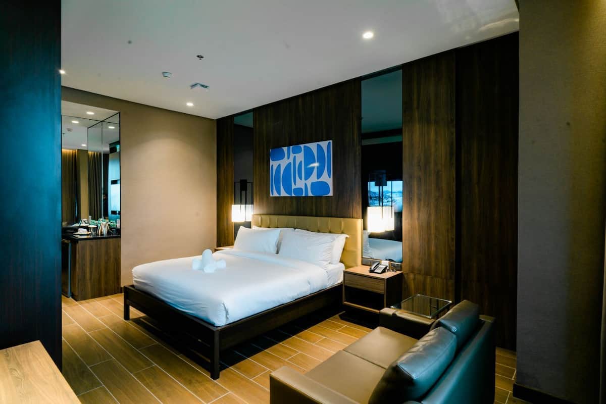 A modern hotel room with a large bed, bedside tables, a seating area, and a view of the city.