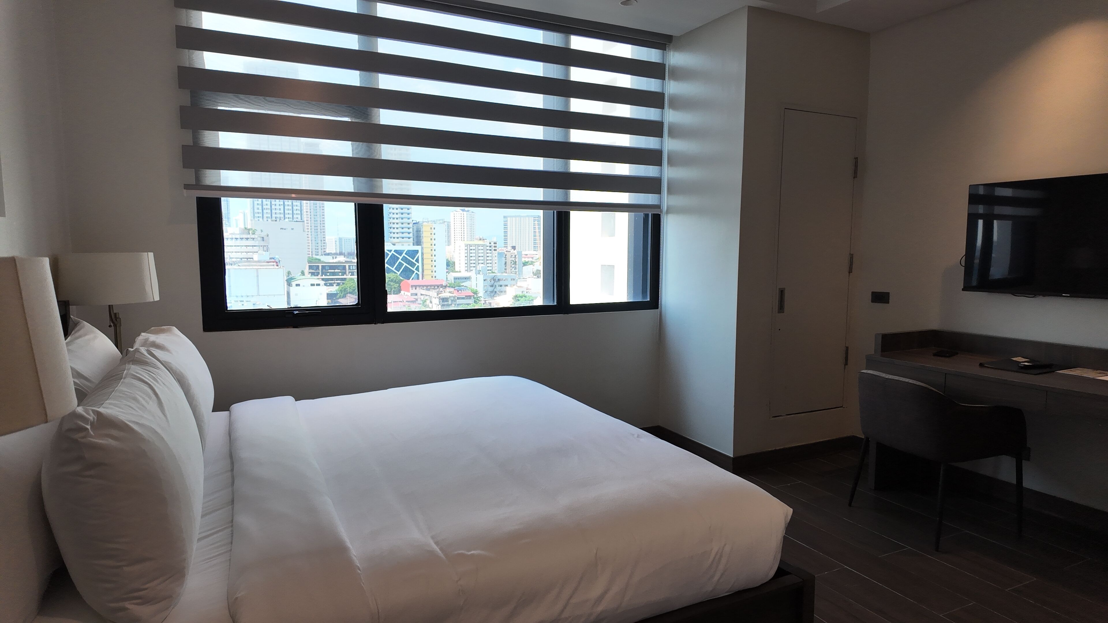 A hotel room with a bed, a desk, a chair, a TV, and a window with blinds.