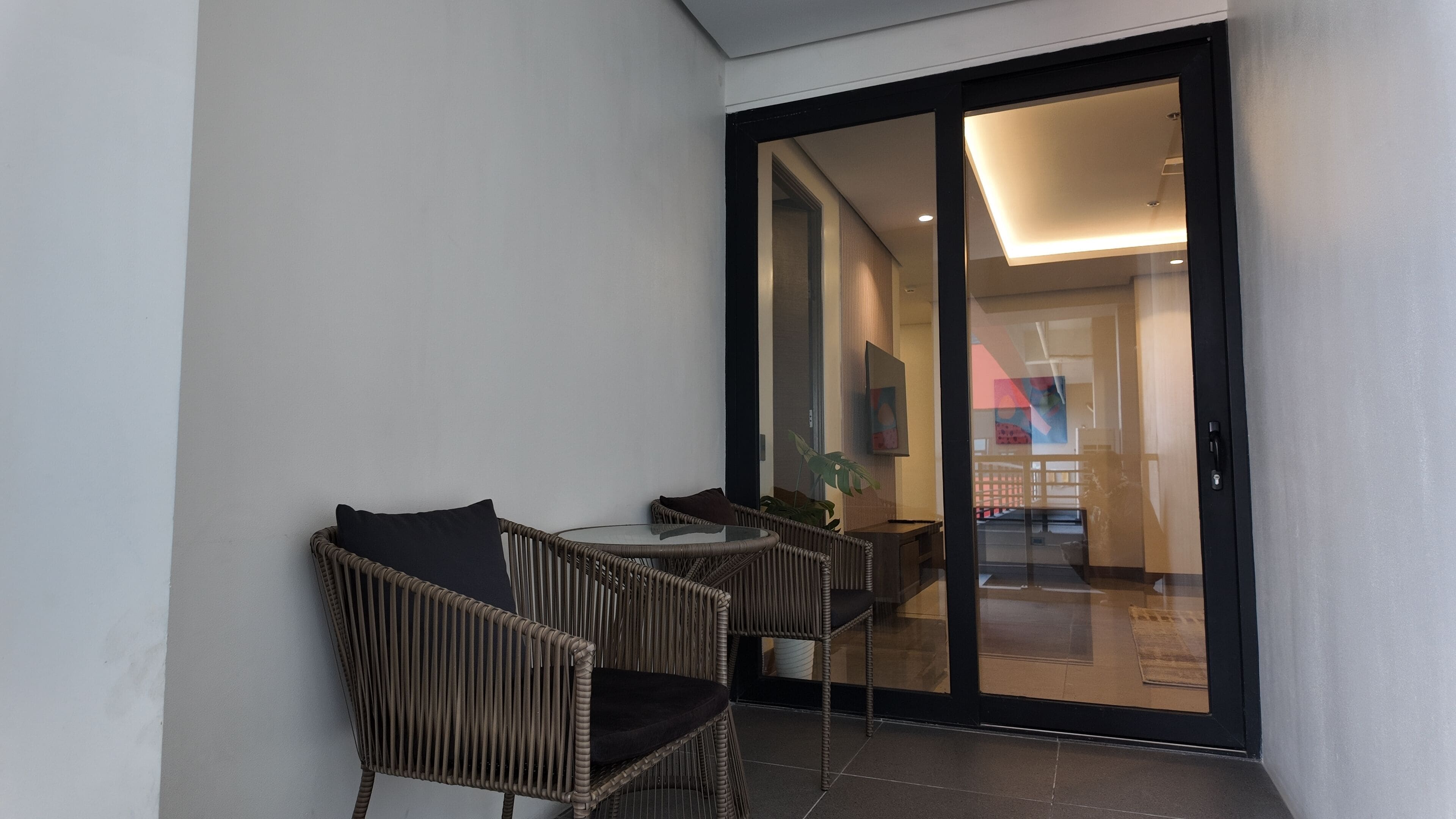 A balcony with wicker chairs and a table, a sliding glass door leading to an interior space with a view of a building's exterior.