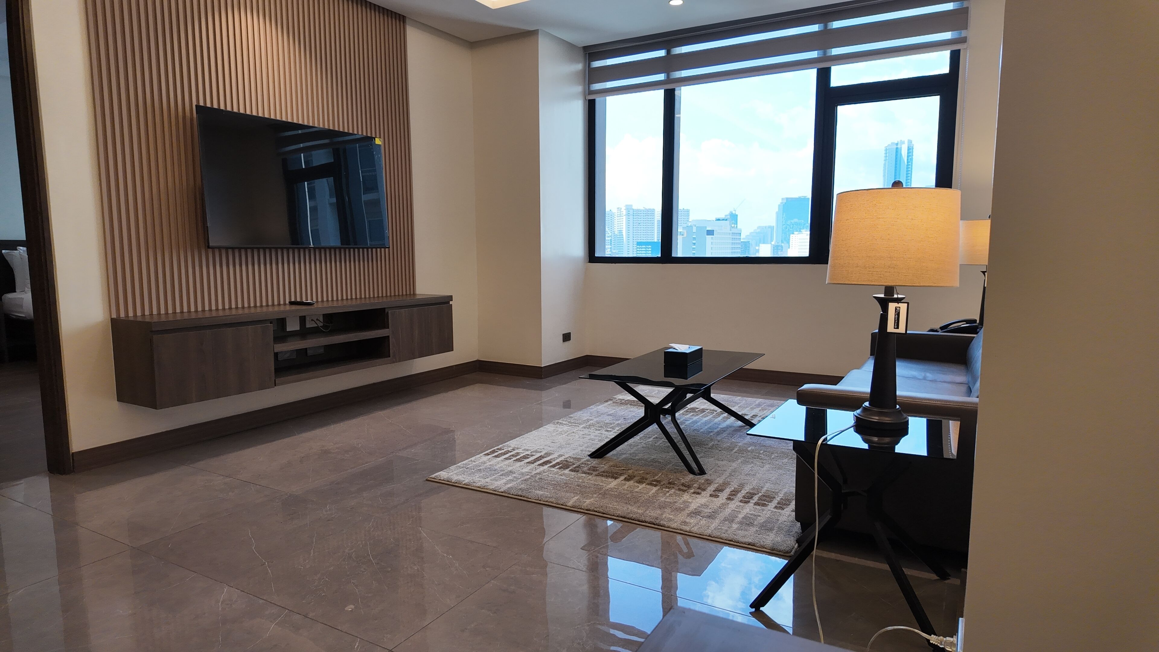 Una sala de estar moderna con un televisor de pantalla plana montado en una pared de paneles de madera, una mesa de centro de vidrio, una lámpara y vistas de la ciudad a través de amplias ventanas.