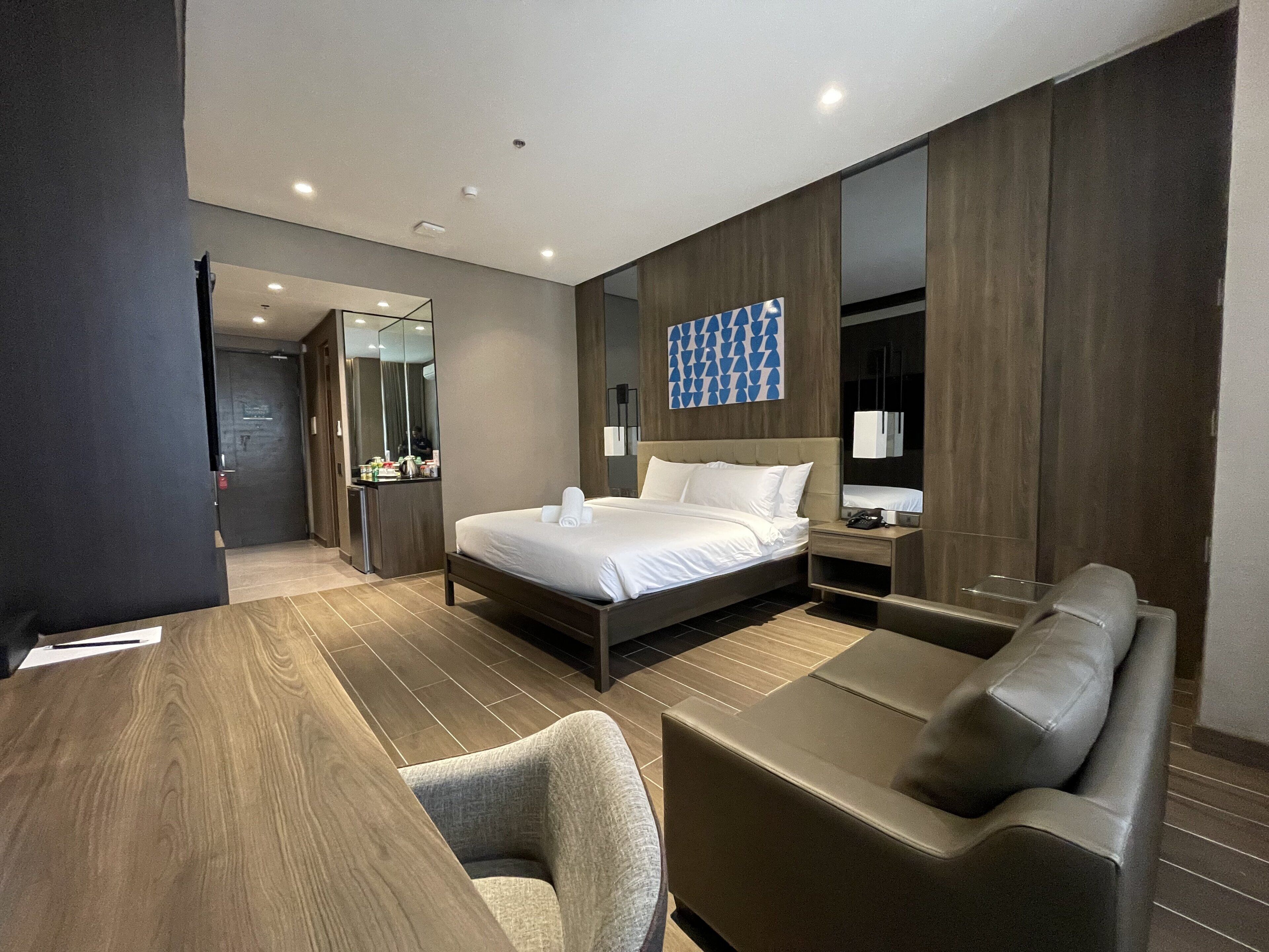 A modern hotel room with a large bed, a desk, a sofa, and a bathroom visible through an open door.