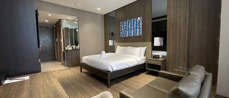 A modern hotel room with a large bed, a desk, a sofa, and a bathroom visible through an open door.