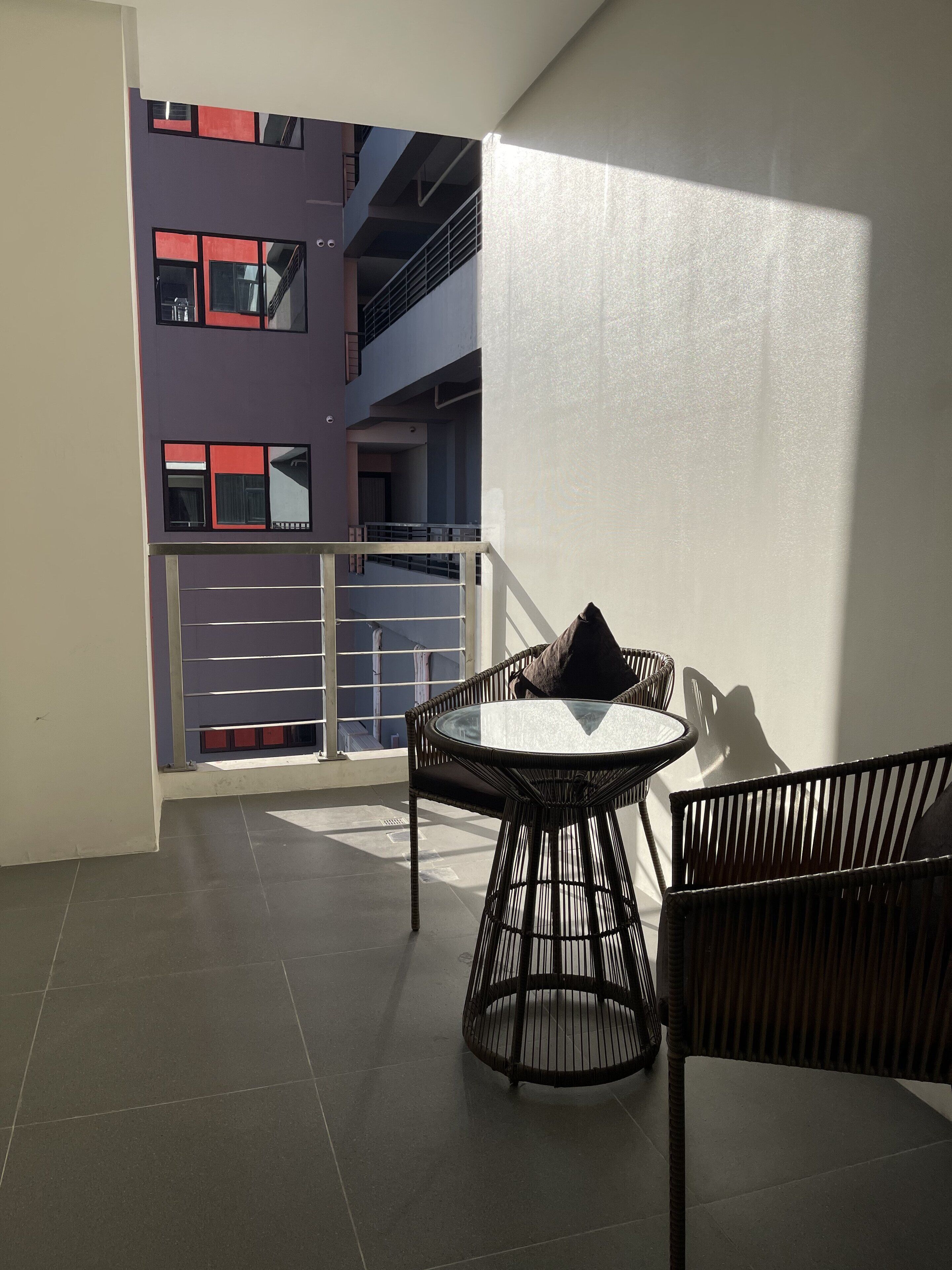 Un balcón con una mesa redonda y sillas de mimbre, con vistas a un edificio con balcones y barandillas.