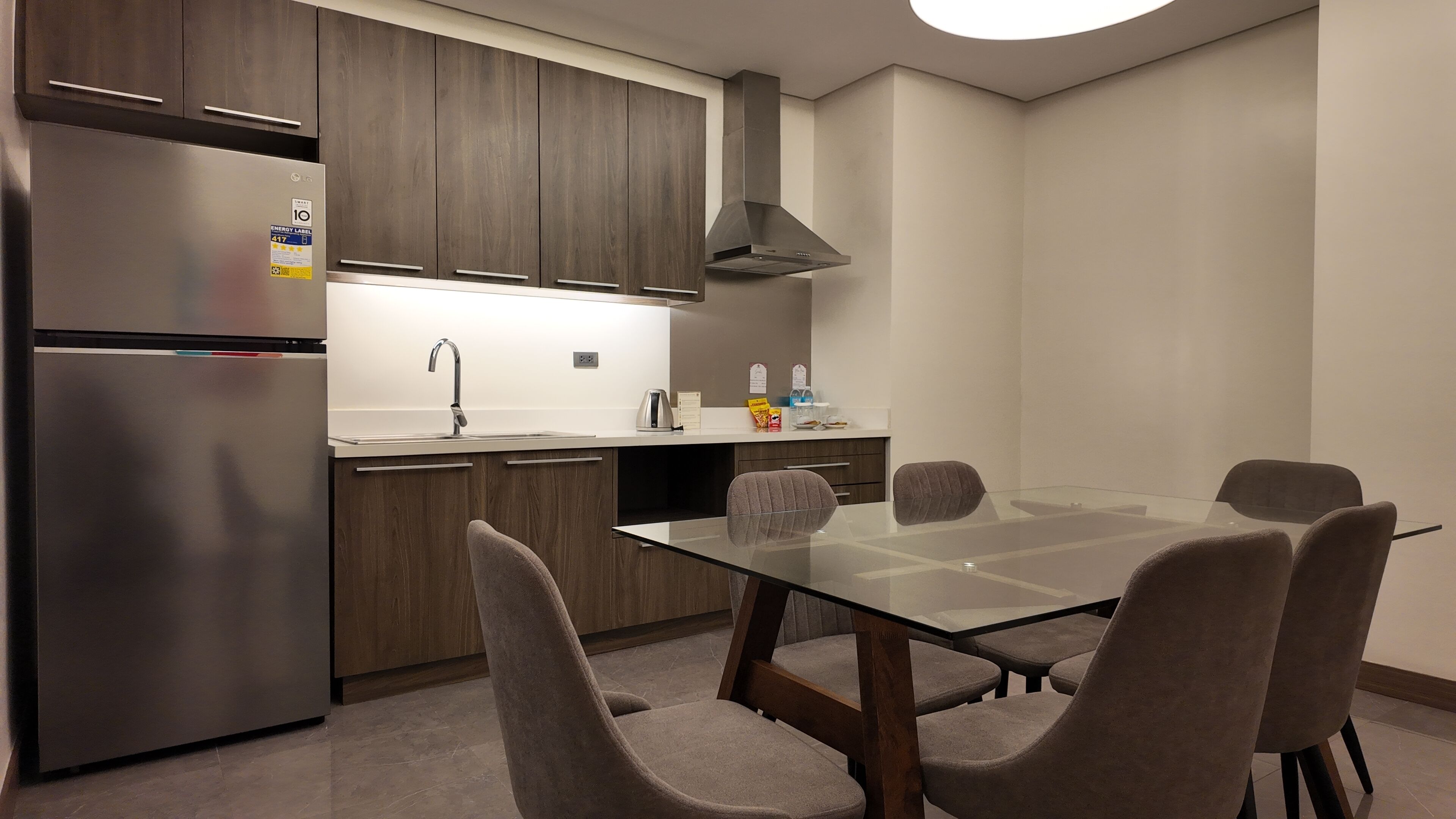A modern kitchen with a stainless steel refrigerator, wooden cabinets, a built-in oven, and a glass dining table with four chairs.
