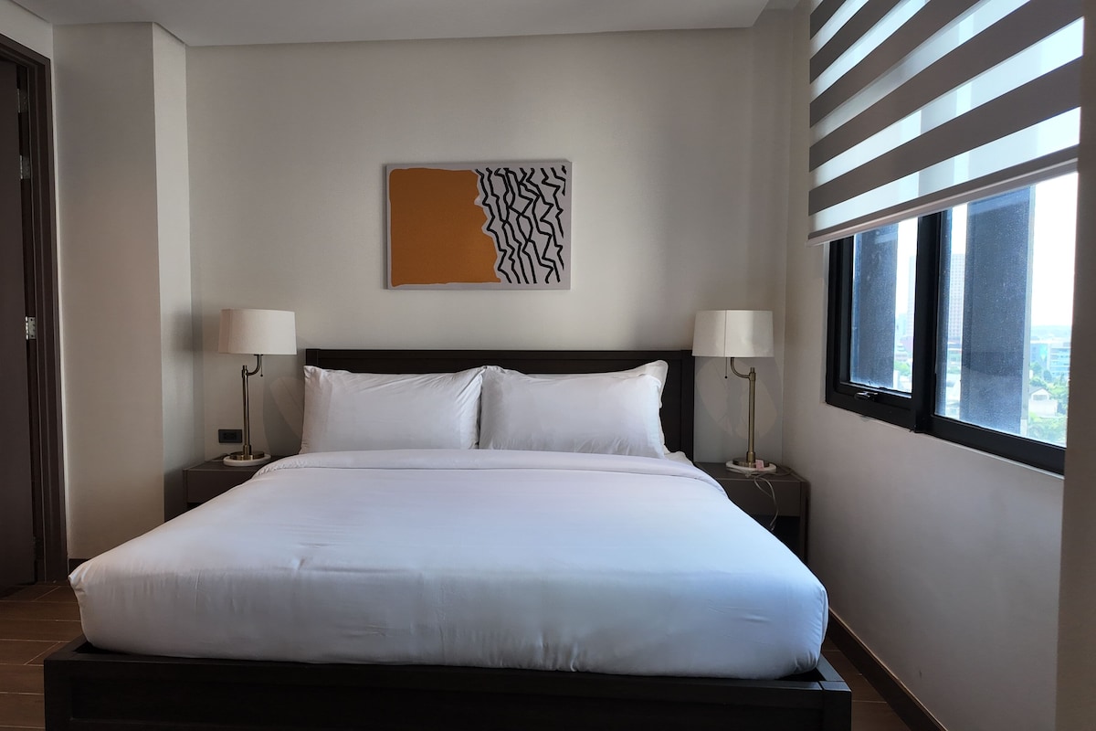 Una habitación de hotel con una cama grande, dos mesitas de noche con lámparas, una ventana con persianas y una obra de arte colgada en la pared.