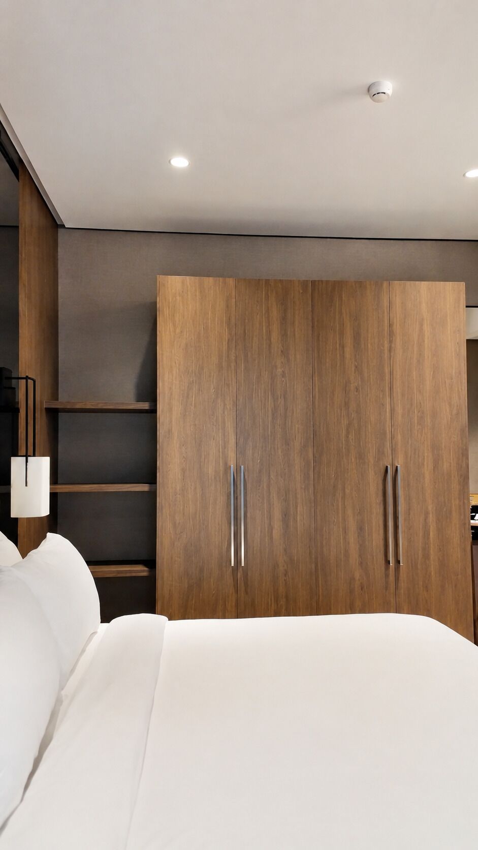 A bedroom with a large wardrobe, a bed with white bedding, and a wall-mounted shelf.