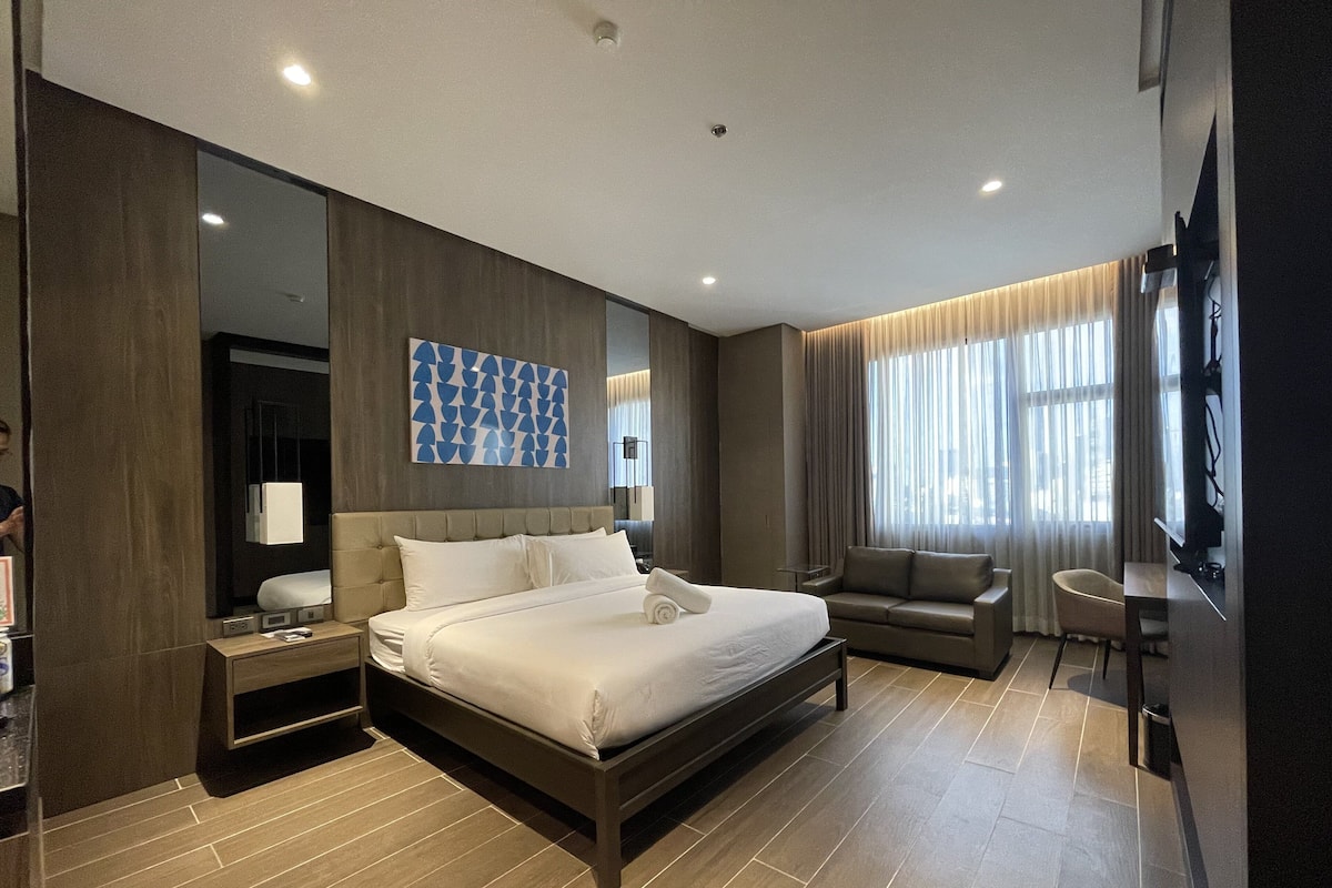 A modern hotel room with a large bed, a sofa, a desk, and a TV.