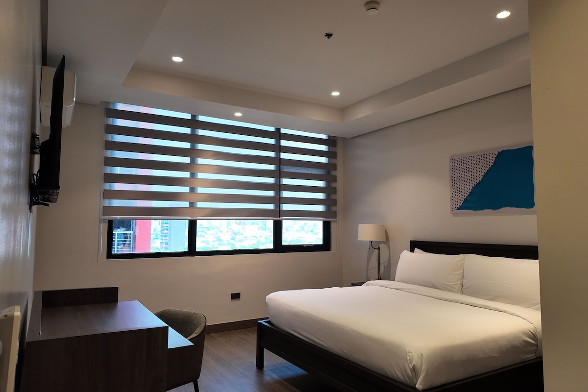Una habitación de hotel con una cama, un escritorio, una silla, un ventanal con persianas, una lámpara de pared y una luz en el techo.