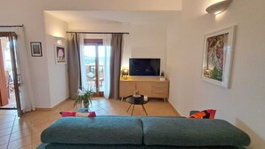 A living room with a sofa, a TV, and a balcony with curtains. - La Terrazza Del Faro - Stunning 1 bedroom flat 600m from the beach (Pittulongu)