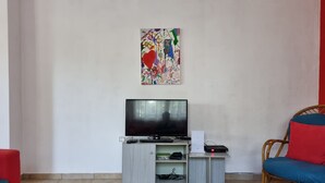 Un salón con un televisor de pantalla plana sobre un mueble bajo, una mesita con un control remoto, un sillón de mimbre con un cojín rojo y un cuadro abstracto y colorido en la pared.